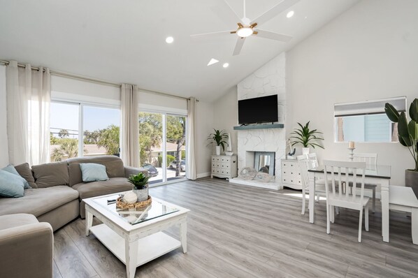 A spacious living room with a large window, a fireplace, a ceiling fan, and a dining area with white furniture.