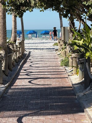 A brick pathway leading to beach umbrellas and chairs, surrounded by palm trees and wooden posts.