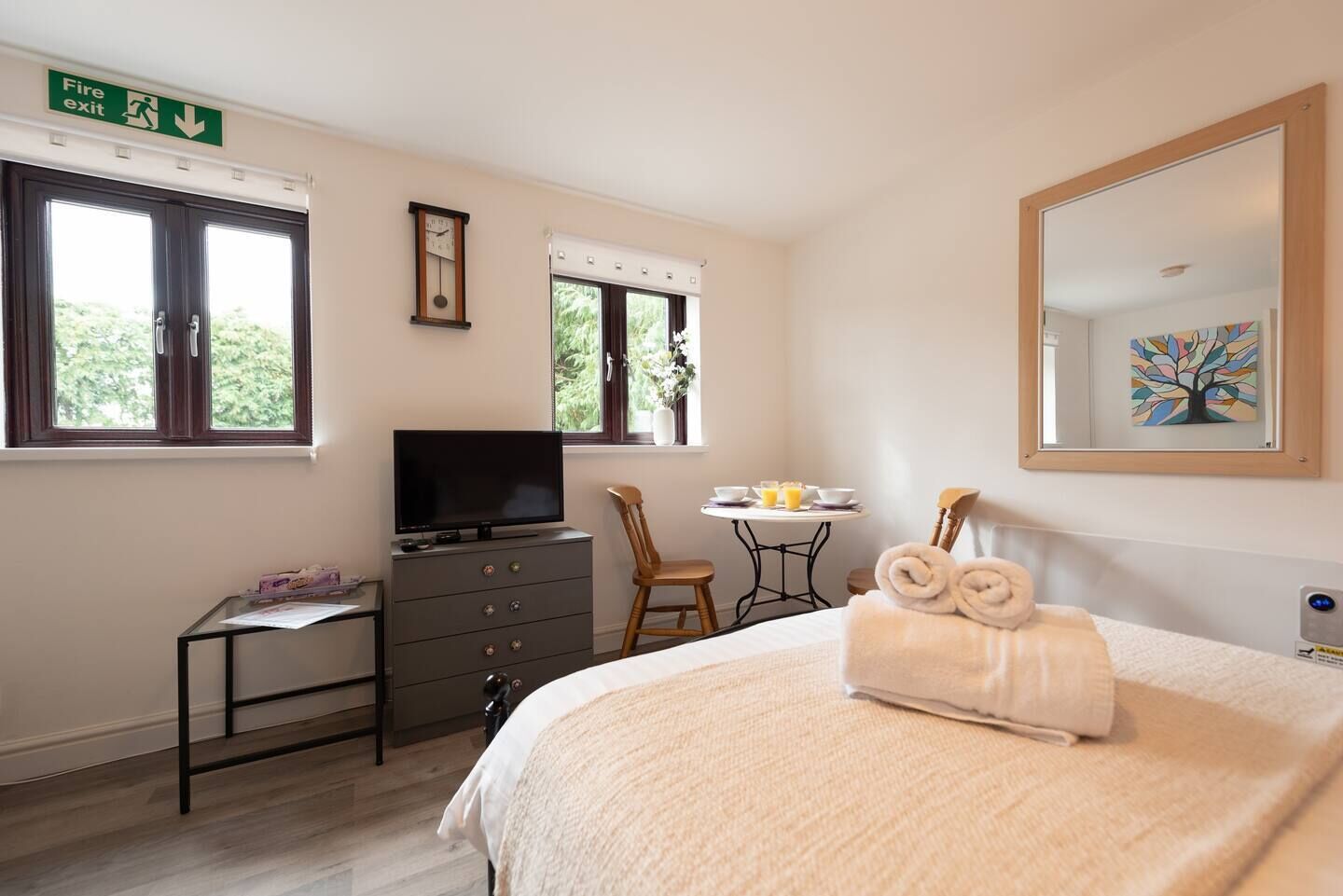 A hotel room with a bed, a television on a dresser, a small table with breakfast items, and a window with a view of greenery.