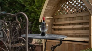 A round metal table with a bottle of wine and a glass on it, set outdoors with a wooden pergola in the background.
