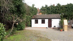 A small white house with a dark brown door and windows, surrounded by a gravel driveway and greenery.