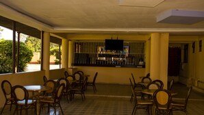 Een restaurant met meerdere tafels en stoelen, een bar met flessen en grote ramen met uitzicht op groen buiten.