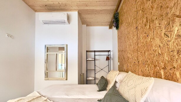 Quarto com teto de madeira, cama com roupa de cama branca, um espelho grande e uma estante de metal.