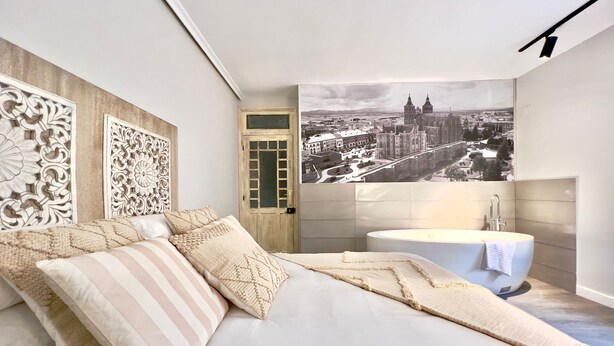 A modern bedroom with a large bed, decorative wall panels, a cityscape mural, and a modern bathroom with a freestanding bathtub. - Espacioso Apartamento con Bañera (Astorga)