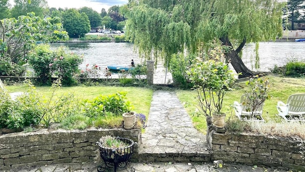 A riverside garden with a stone pathway, seating area, and a large willow tree. - Lux 8Br Thamesfront Villa w/ Yacht & Sauna (Staines)