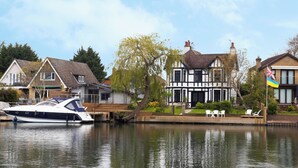 A waterfront property with a boat docked, a large house with a thatched roof, and a flag. - Lux 8Br Thamesfront Villa w/ Yacht & Sauna (Staines)