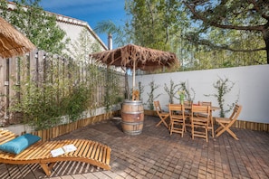 Eine Terrasse im Freien mit einem Holztisch, Stühlen, einer Holzbank und einem reetgedeckten Sonnenschirm.