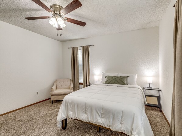 A bedroom with a bed, a chair, a nightstand, a ceiling fan, and a window with curtains.
