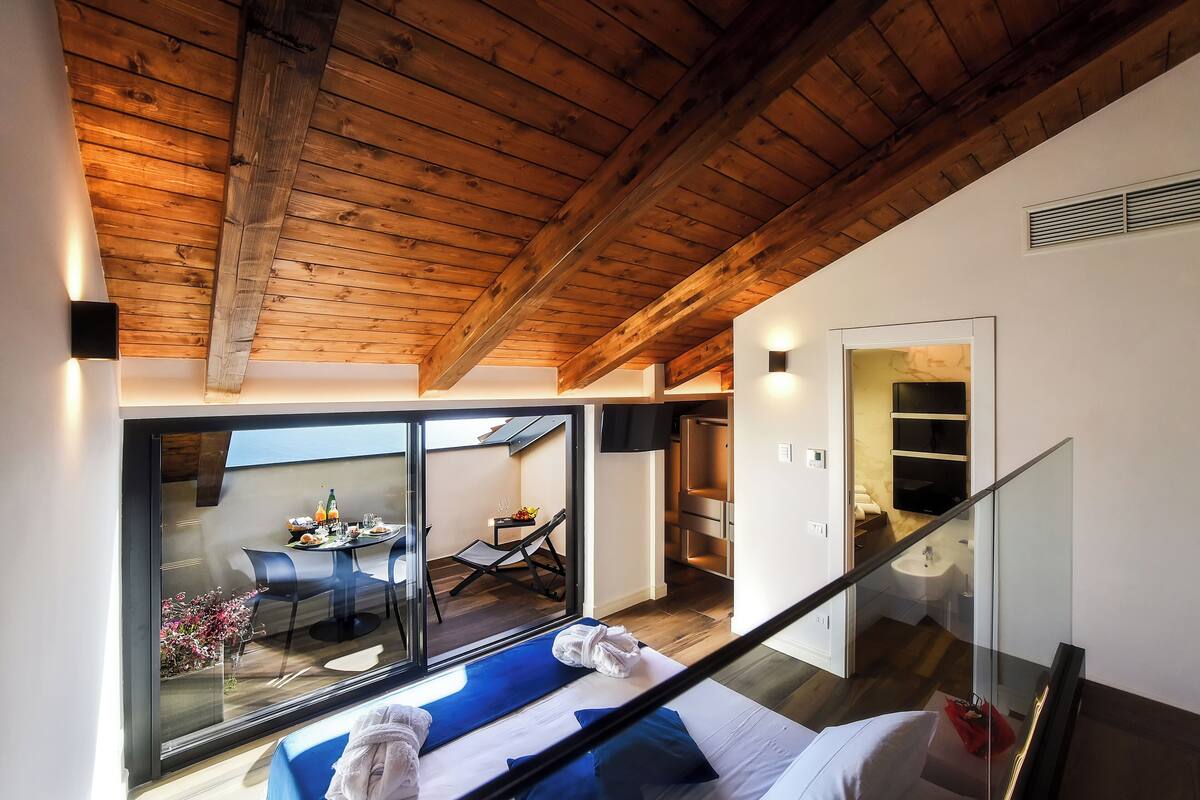 Habitació d'hotel moderna amb sostre de fusta, un balcó amb zona de menjador i una porta de vidre que dona a un bany.