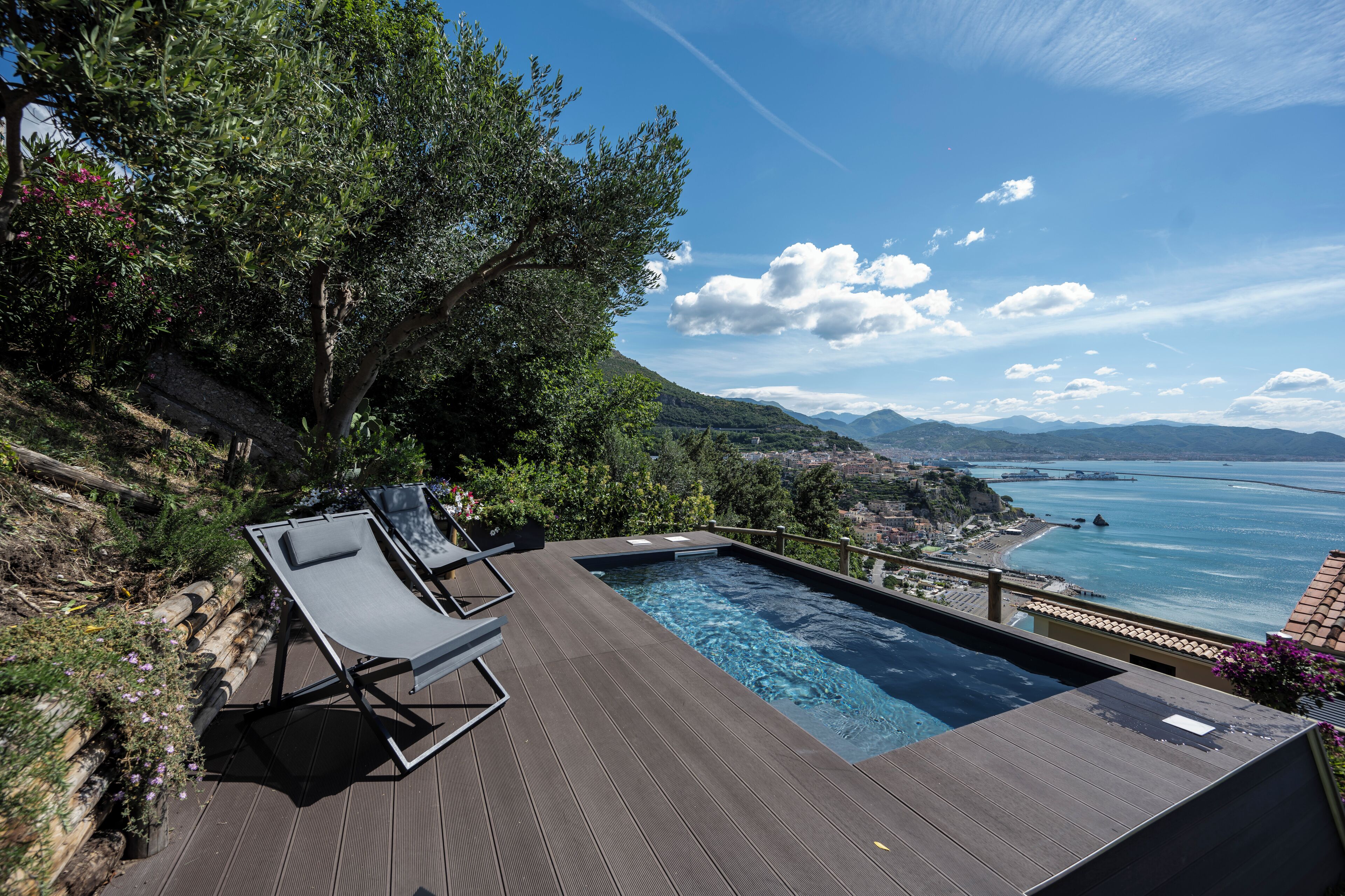 A swimming pool with lounge chairs on a wooden deck, overlooking a coastal landscape with mountains and a bridge.