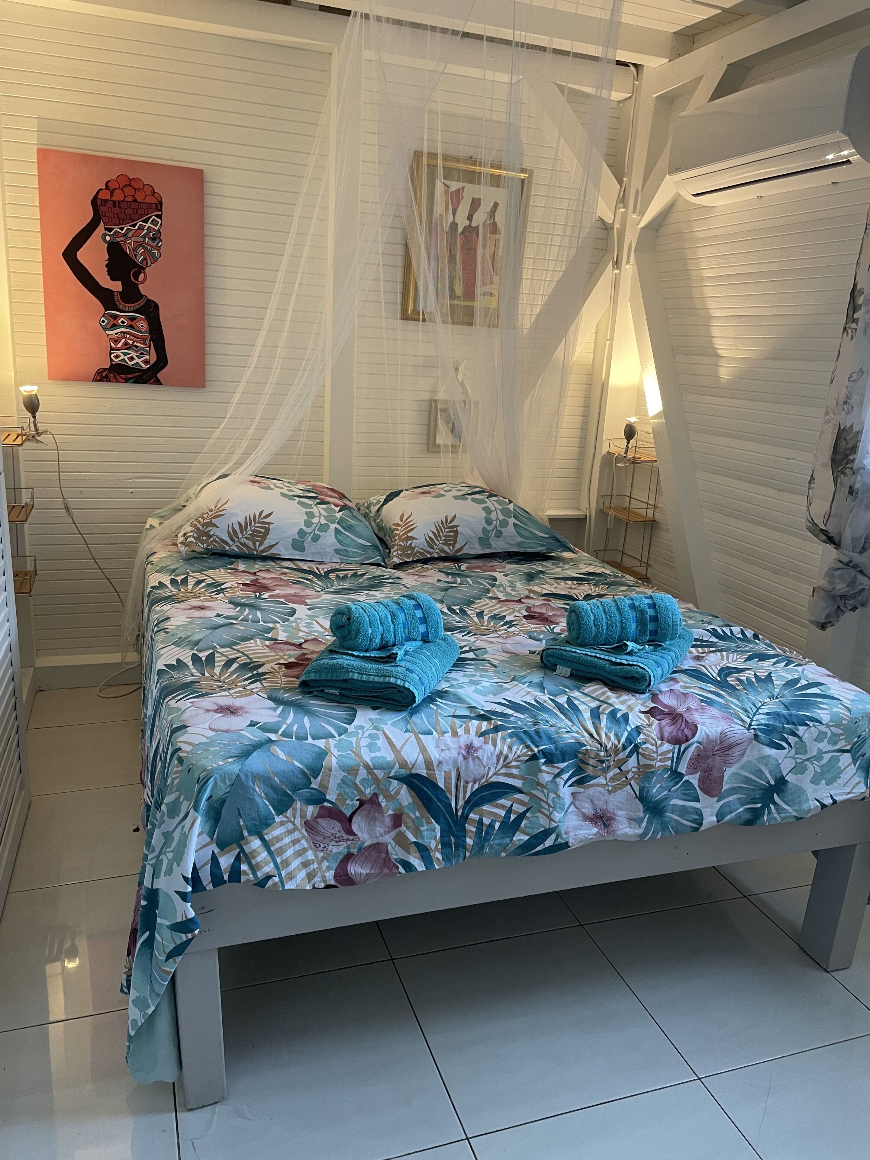 A bed with a floral bedspread and blue towels, a white headboard, and artwork on the wall.