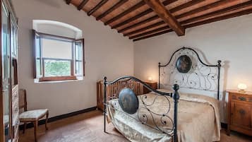 A bedroom with a wooden ceiling, a bed with a metal headboard, a window with a view, a wooden nightstand, and a chair.