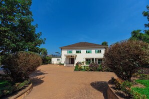 A two-story white building with a brown roof, surrounded by well-maintained greenery and a paved driveway. - Family-sized unique private villa w/an office & kitchen, super close to the city (Kampala)