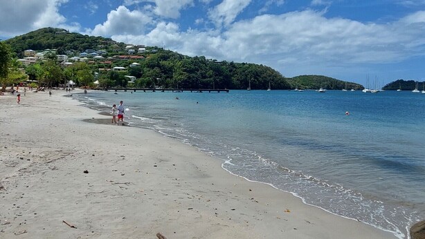 A beach with clear blue water, a sandy shore, and a hill with buildings in the background.