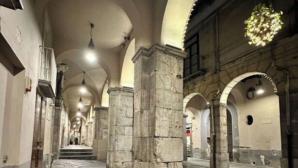 Un pasillo con columnas de piedra, techos abovedados y lámparas colgantes.