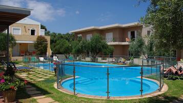 A swimming pool with a surrounding deck, a covered seating area, and a building with balconies in the background.