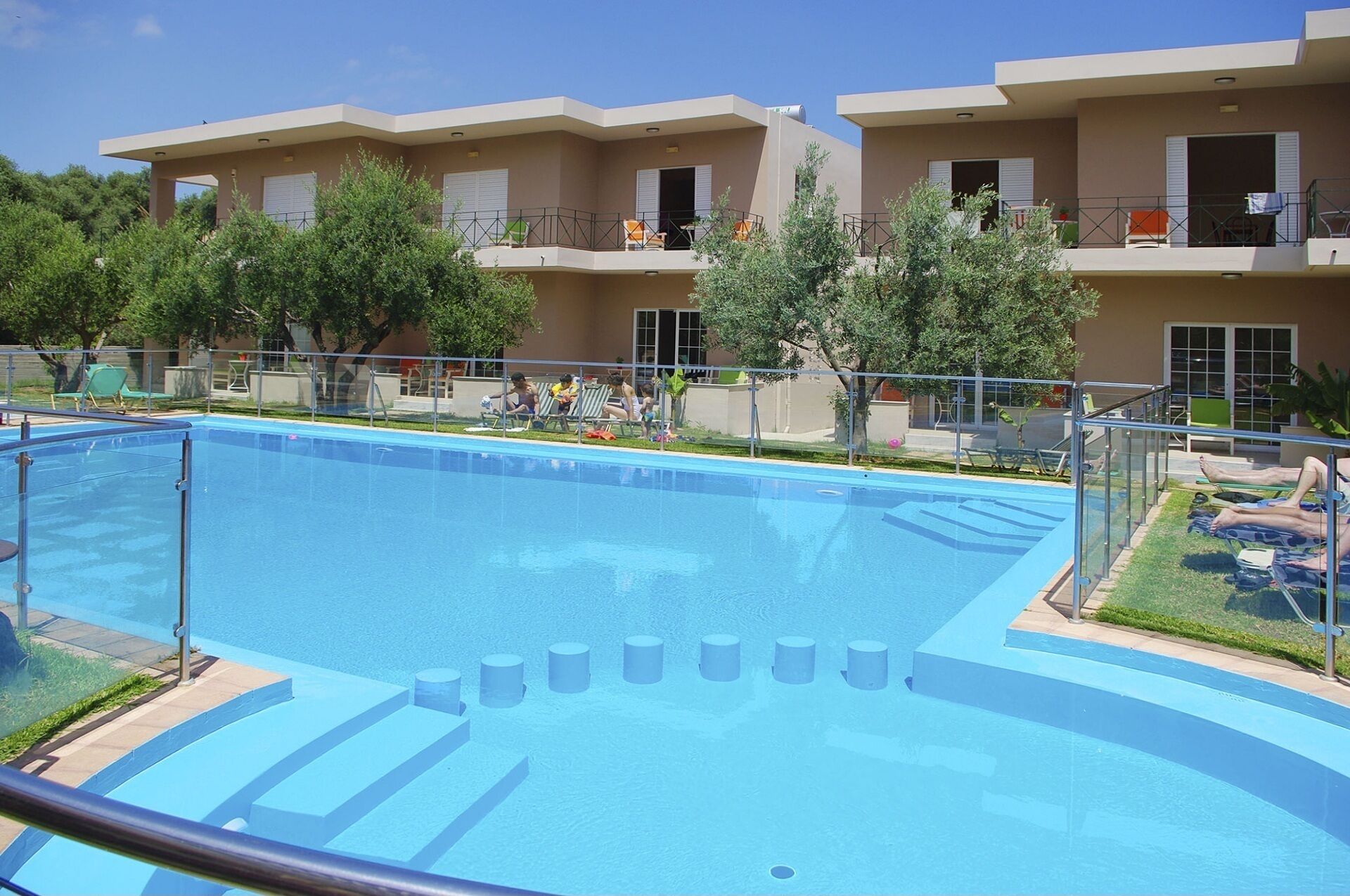Une piscine avec un bassin aux eaux d’un bleu limpide, entourée d’une petite clôture. On y trouve des chaises longues et un petit espace aménagé avec vue sur un immeuble à plusieurs étages doté de balcons.