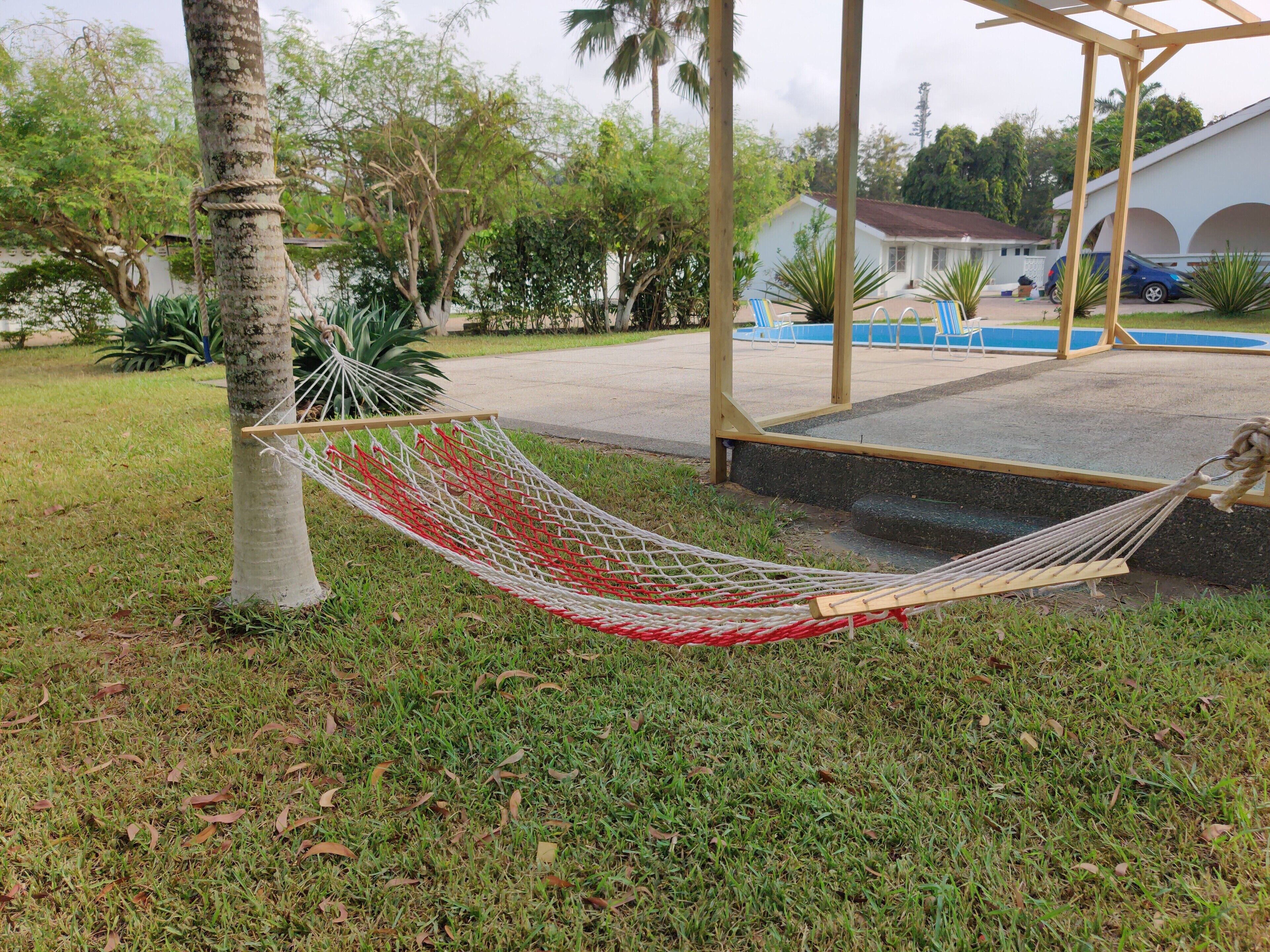 A hammock tied to a tree in a grassy area with a pool and a covered patio in the background.