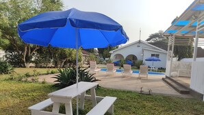 A patio area with a white picnic table and a blue umbrella, surrounded by white chairs and a pool.