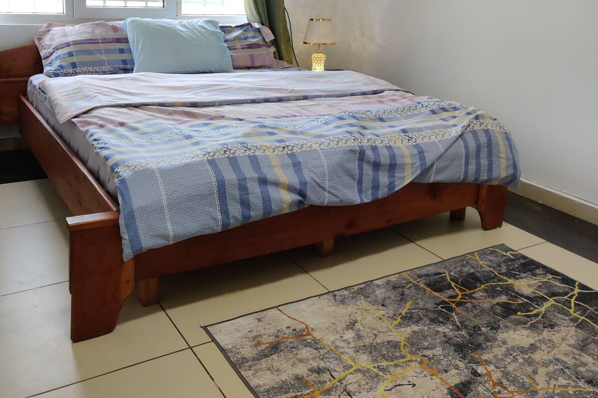 Un dormitori amb llit de fusta, una taula de nit, una làmpada, una finestra amb cortines i una catifes amb dibuixos.