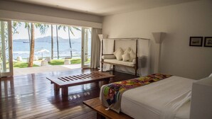 A bedroom with a bed, a wooden bench, and a daybed with pillows. There is a large window with a view of the ocean and palm trees. - Coconut Palms and Endless Blue Skies! (Candidasa)