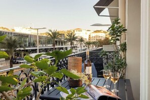 A balcony with a table, a bottle, and two glasses of wine, overlooking a cityscape with buildings and palm trees.