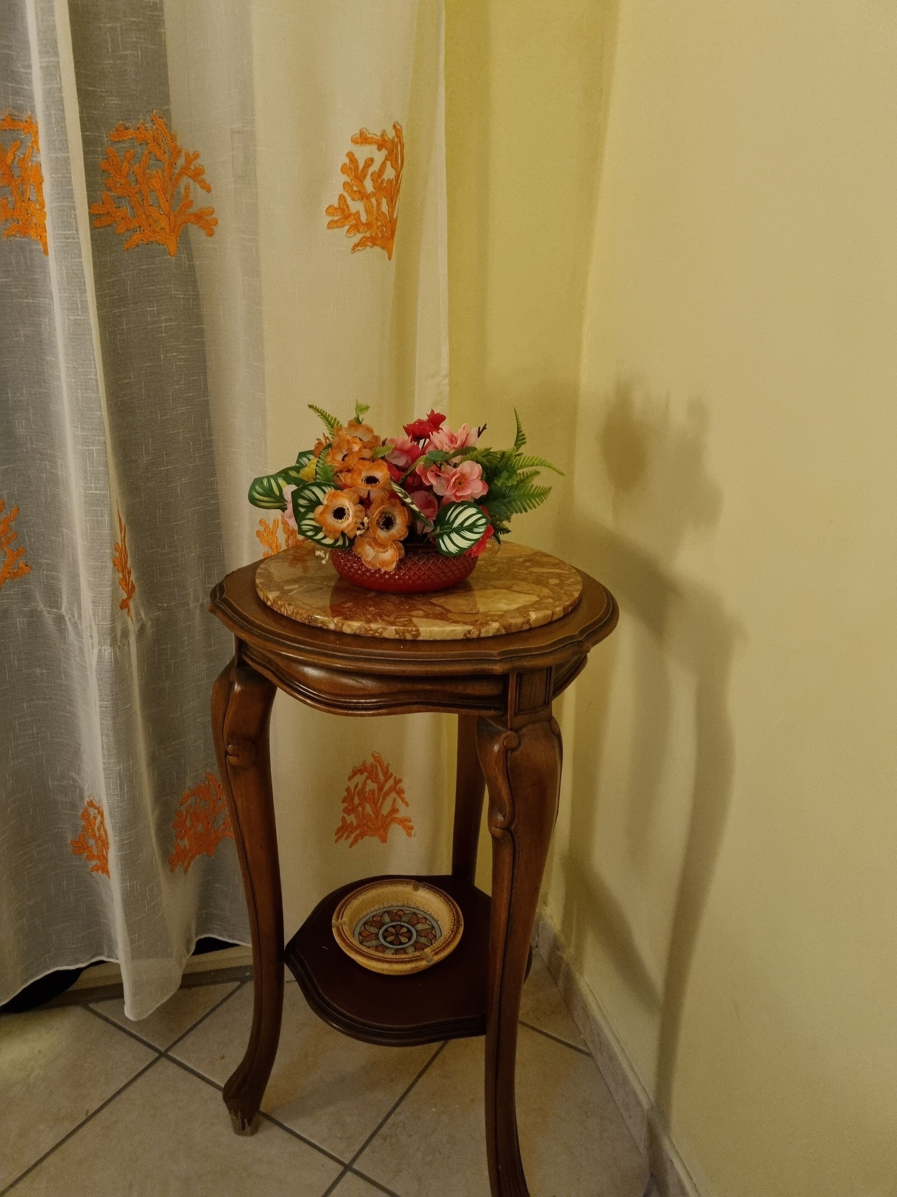 A wooden table with a decorative bowl containing flowers, positioned in a corner with a curtain featuring orange leaf patterns.