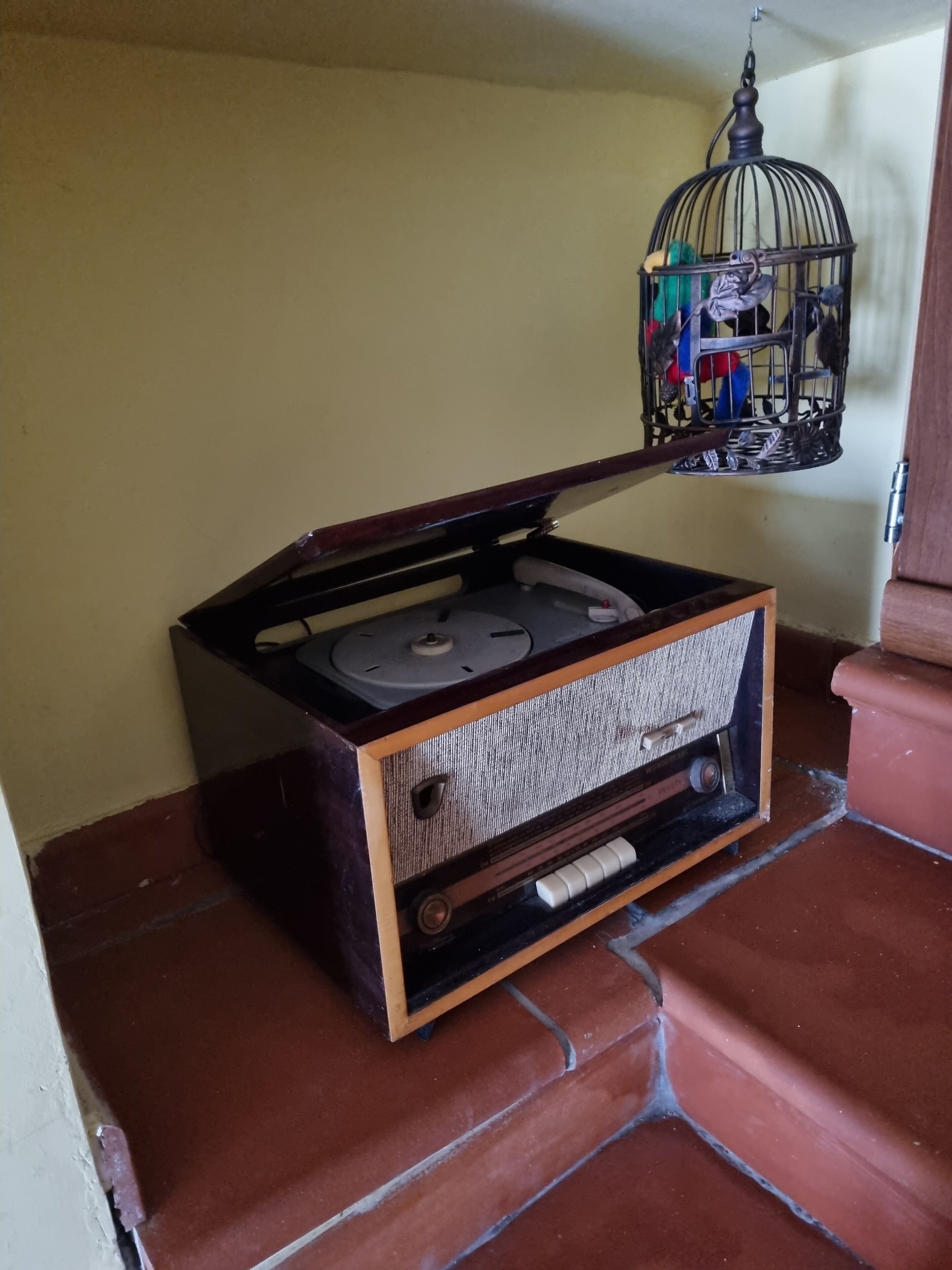 Une radio ancienne, avec un boîtier en bois et une grille en métal, posée sur un sol carrelé, près d’un mur.