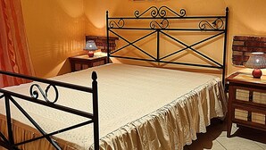 Una cama con estructura metálica y una colcha de encaje, una mesita de noche de madera con una lámpara, y una pared de ladrillo con un cuadro.