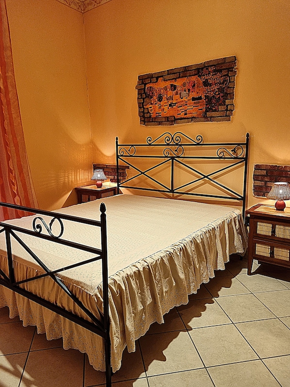 A bed with a metal frame and a lace bedspread, a wooden nightstand with a lamp, and a brick wall with a painting.