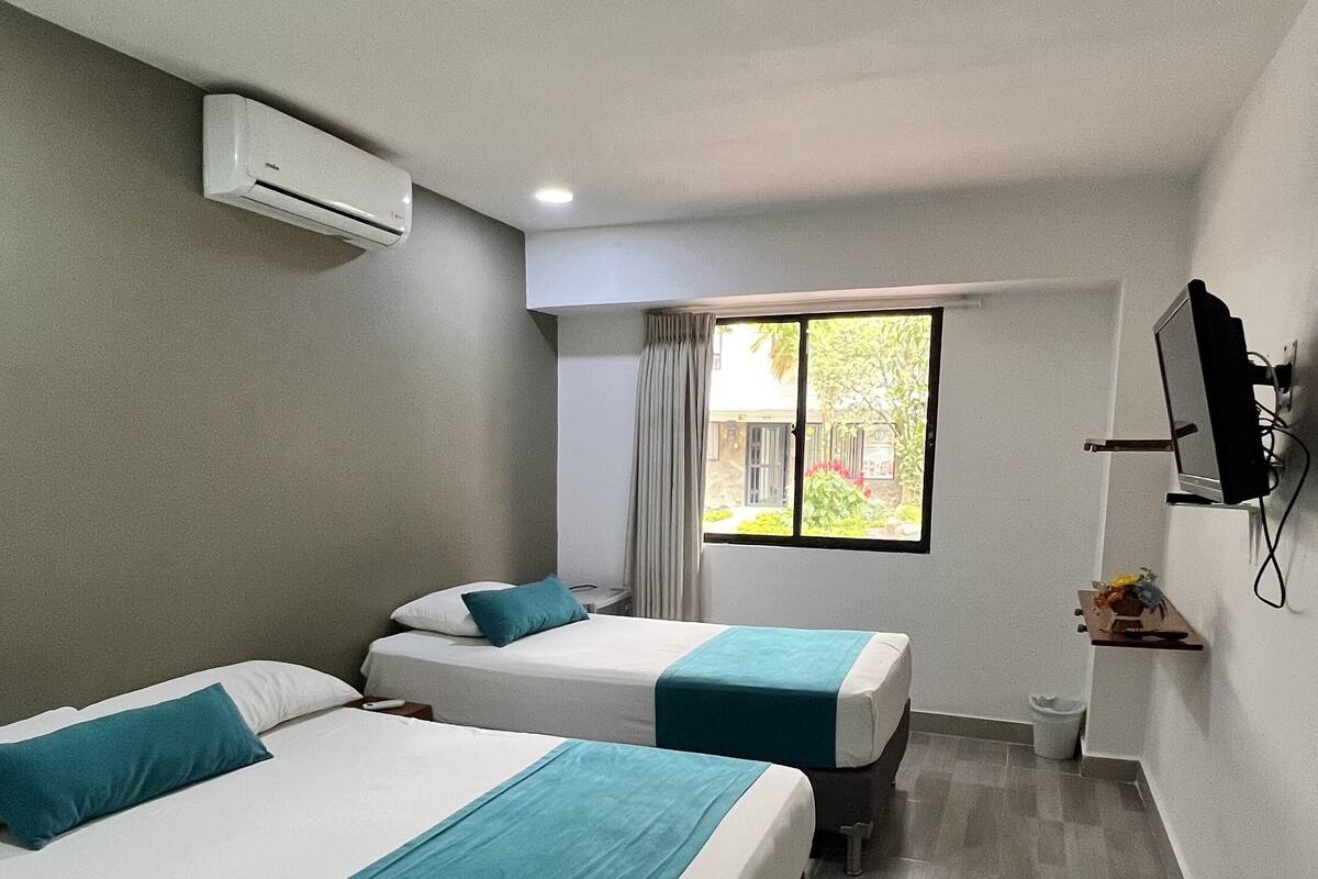A hotel room with two beds, a window with curtains, an air conditioning unit, a small table, and a TV mounted on the wall.