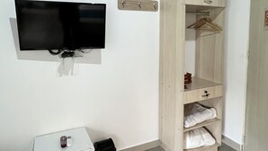 Una habitación con una televisión de pantalla plana montada en la pared, un armario de madera con estantes, un pequeño mueble blanco y un calentador en el suelo.