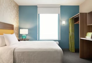 Habitación de hotel con cama, mesita de noche con lámpara, ventana con vistas, armario y escritorio.