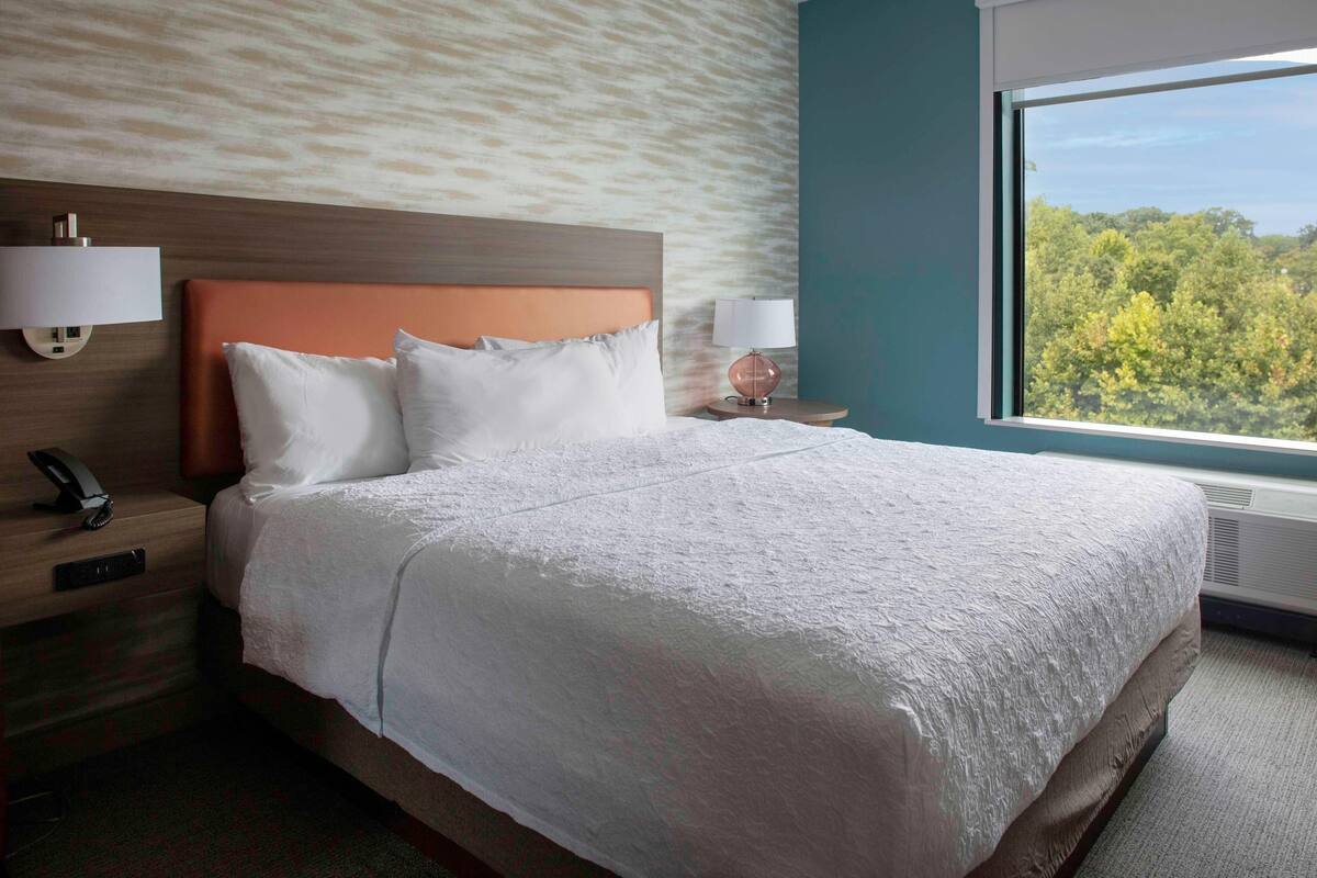 A hotel room with a large bed, two bedside tables with lamps, a wall-mounted phone, and a window with a view of trees.