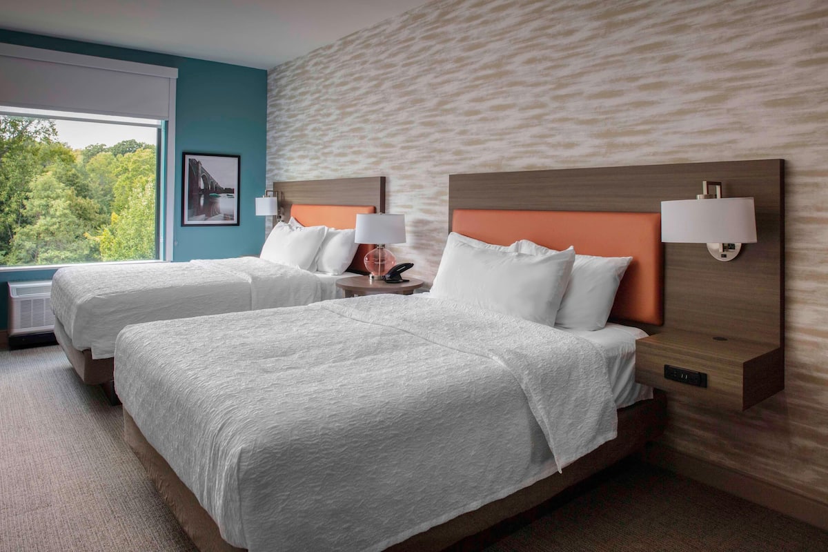 A hotel room with two beds, a large window, and a view of greenery.