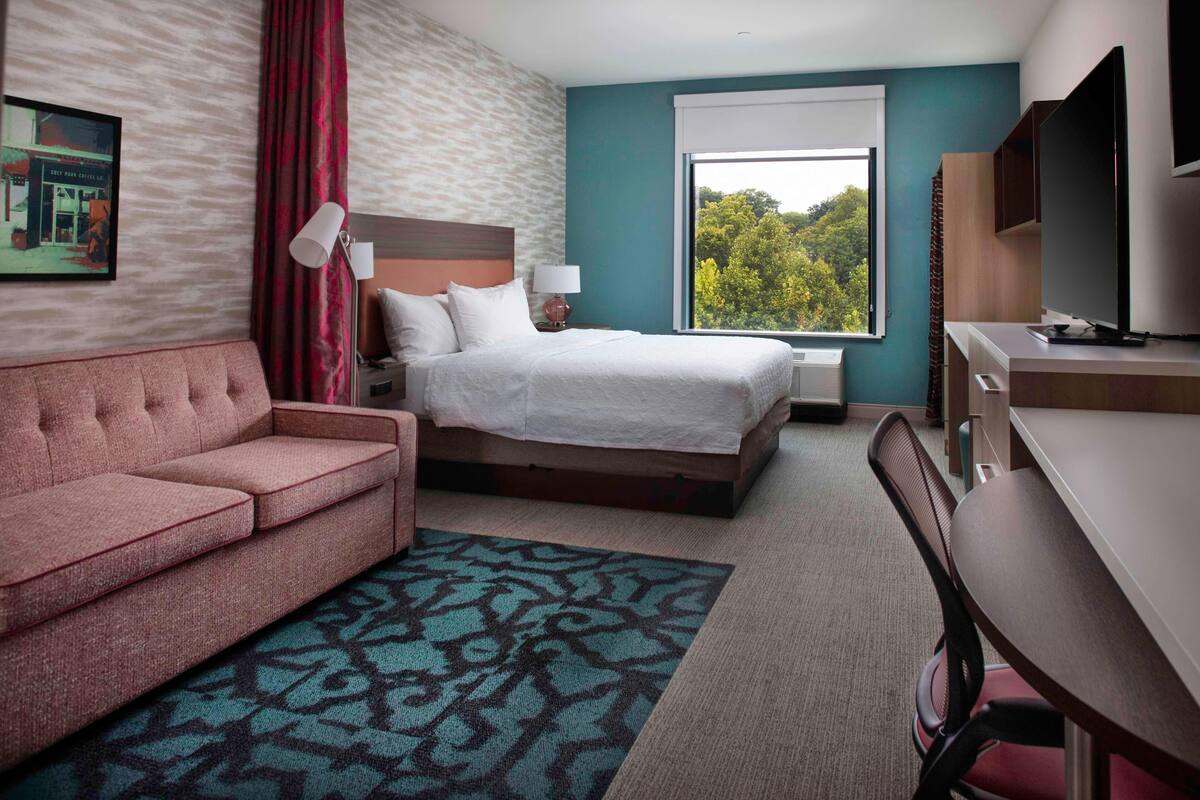 A hotel room with a bed, a desk, a sofa, and a large window offering a view of trees.