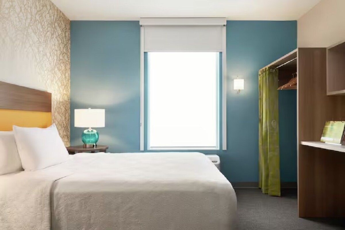 Habitación de hotel con cama, mesita de noche con lámpara, ventana con vistas, armario y escritorio.