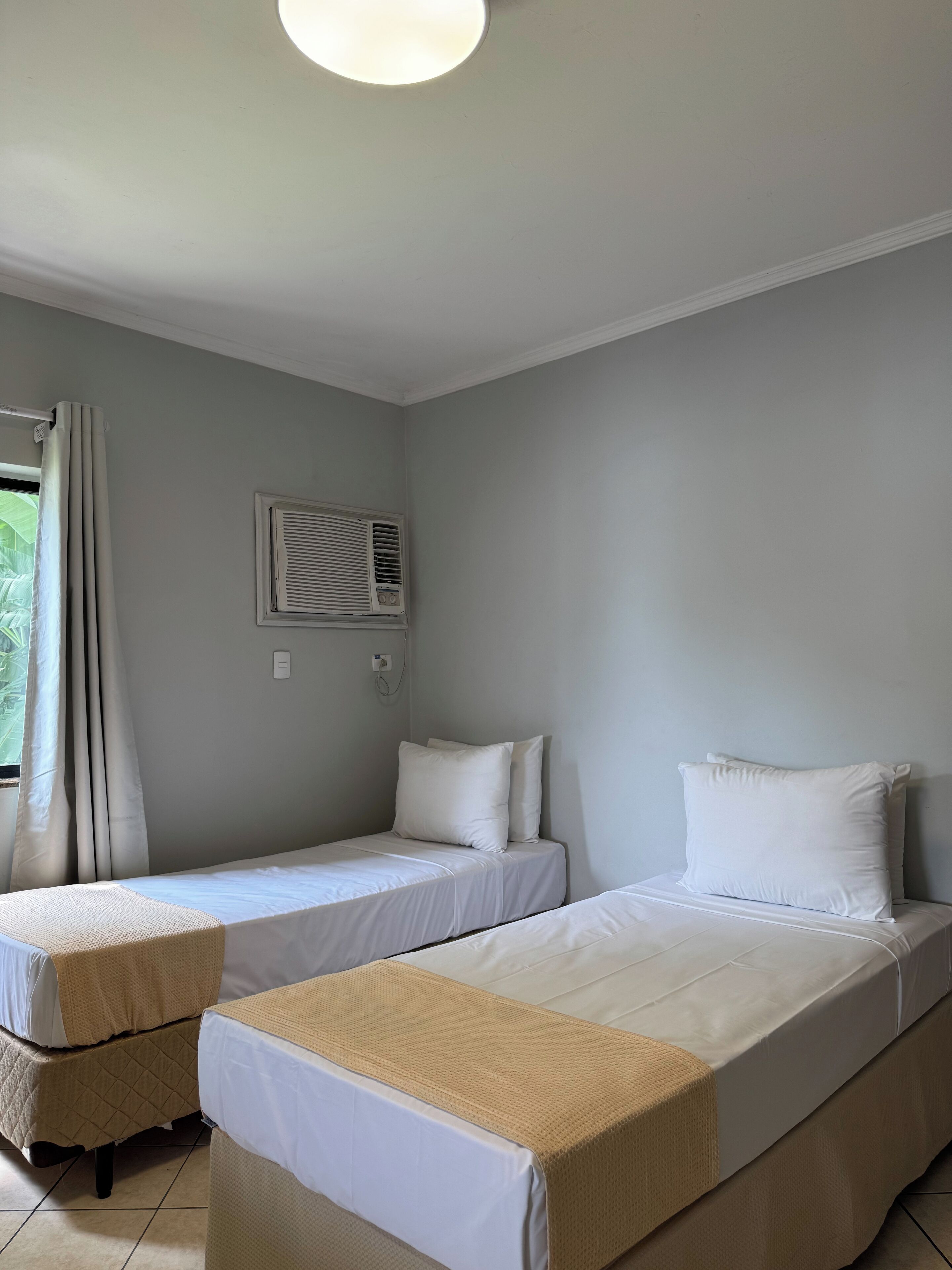Una habitación con dos camas individuales, una ventana con cortinas, un equipo de aire acondicionado y una lámpara fijada a la pared.