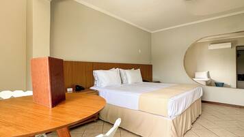 A hotel room with a bed, a wooden table, a white chair, and a small kitchenette area.