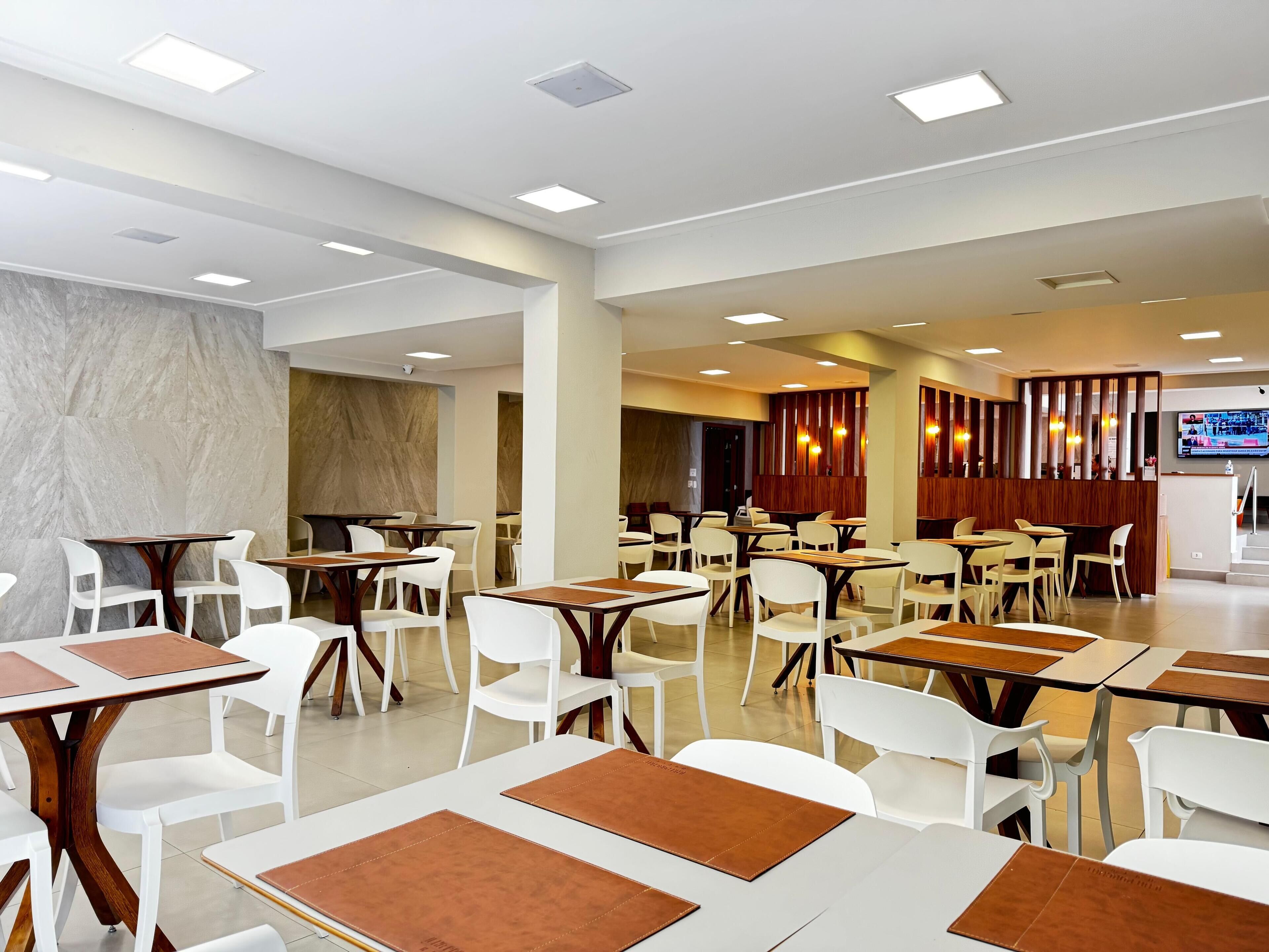 A spacious restaurant with white chairs and tables, recessed ceiling lights, and a TV screen on the wall.