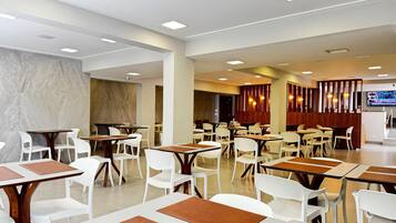 A spacious restaurant with white chairs and tables, recessed ceiling lights, and a TV screen on the wall.