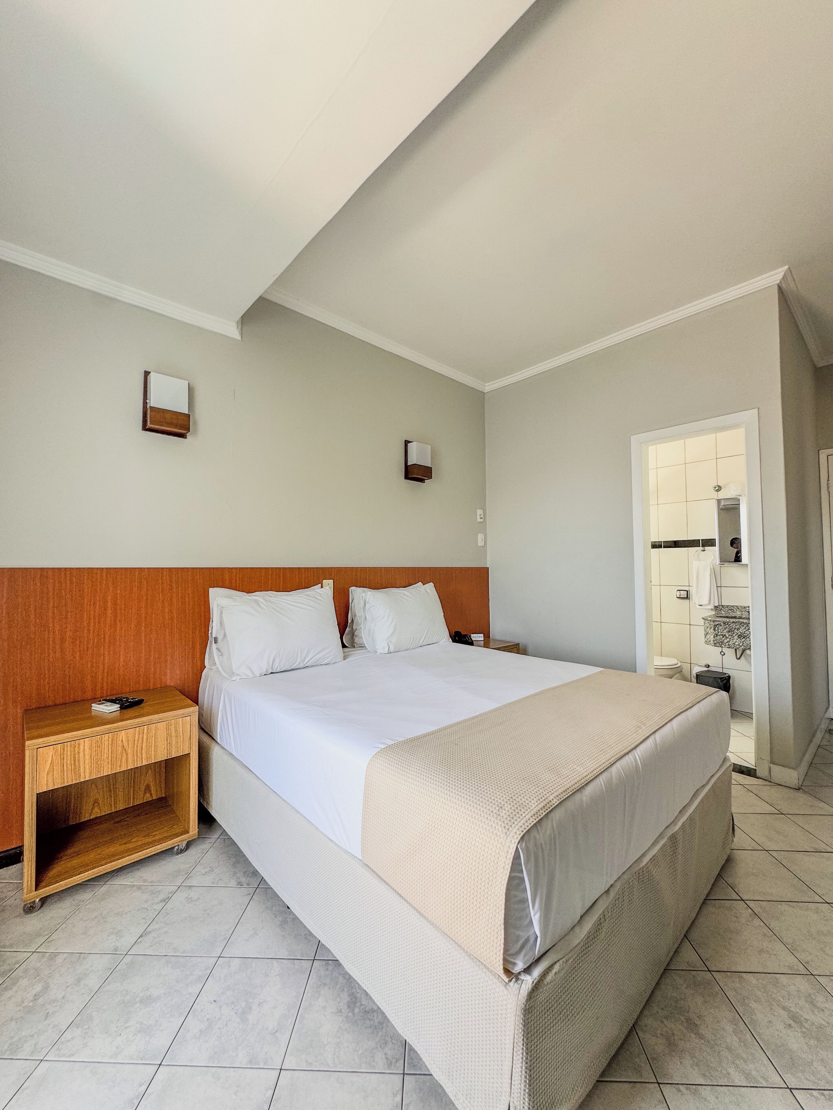 Habitación de hotel con una cama grande, cabecera de madera, dos mesitas de noche, un tocador y un baño visible a través de una puerta abierta, con suelo de baldosas.