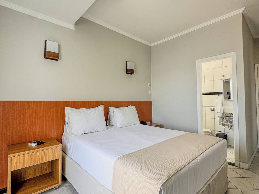 Habitación de hotel con una cama grande, cabecera de madera, dos mesitas de noche, un tocador y un baño visible a través de una puerta abierta, con suelo de baldosas.