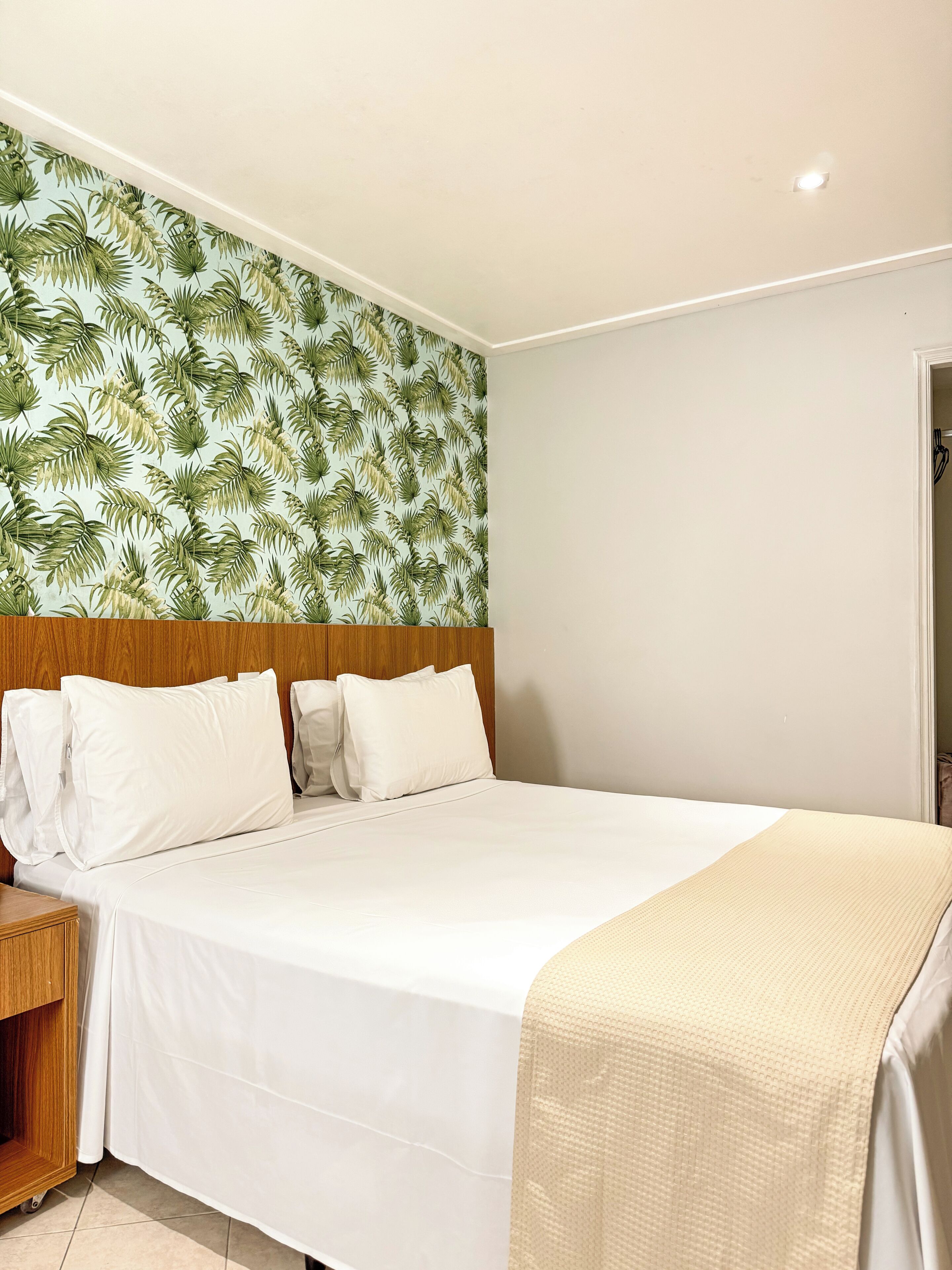 Ein Hotelzimmer mit Bett, Holz-Kopfteil, weißer Bettwäsche und tropischem Blattmuster an der Tapete.