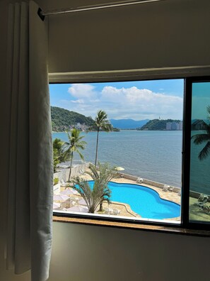 Une vue depuis une chambre dotée d’une grande fenêtre donnant sur une piscine, des palmiers et une étendue d’eau, avec, au loin, des bâtiments et des collines.