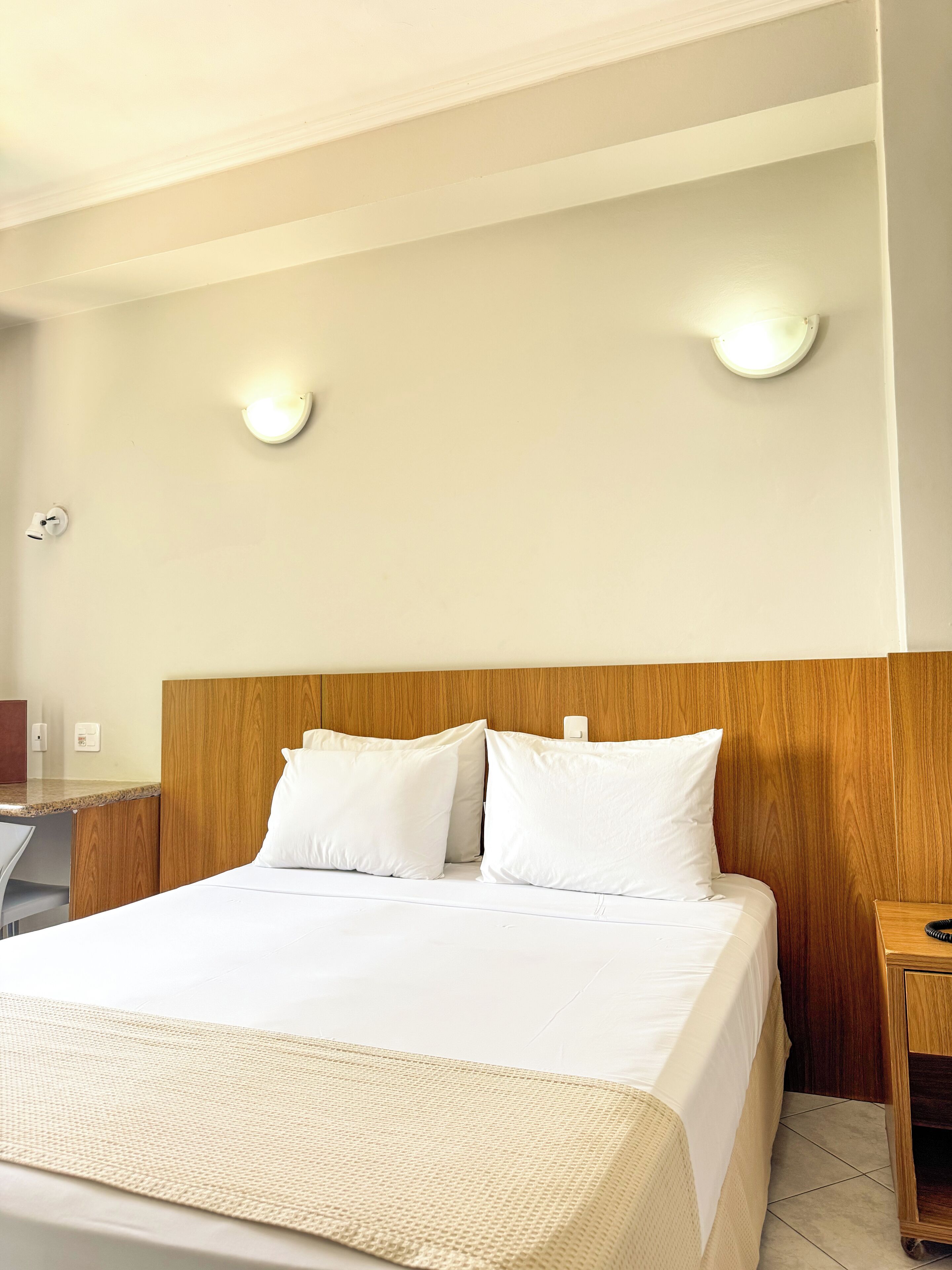 A hotel room with a wooden headboard, a bed with white bedding, a small wooden nightstand, and two wall-mounted lights.