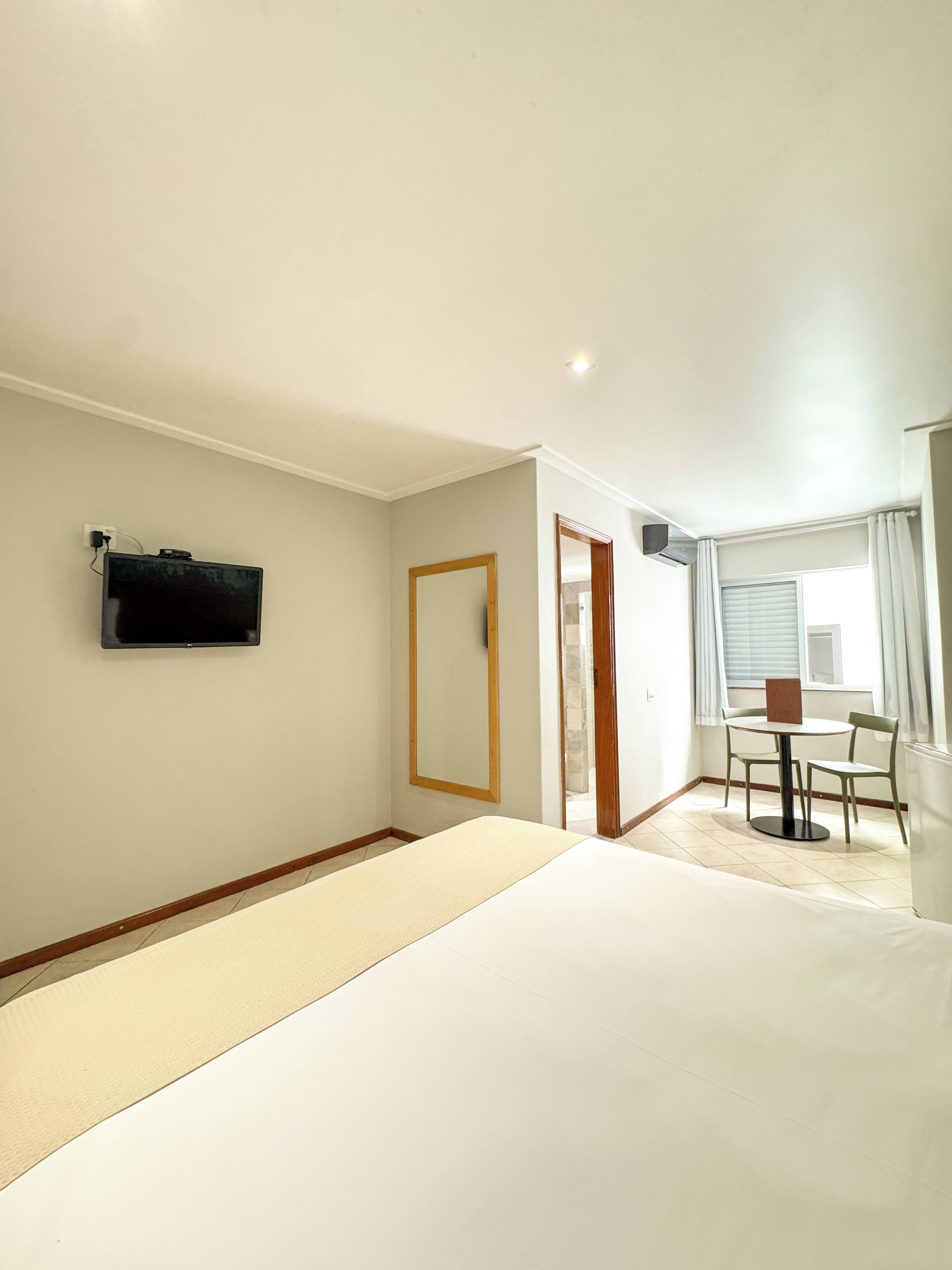 A hotel room with a bed, a TV mounted on the wall, a wooden door, a dining table with chairs, and a window with curtains.