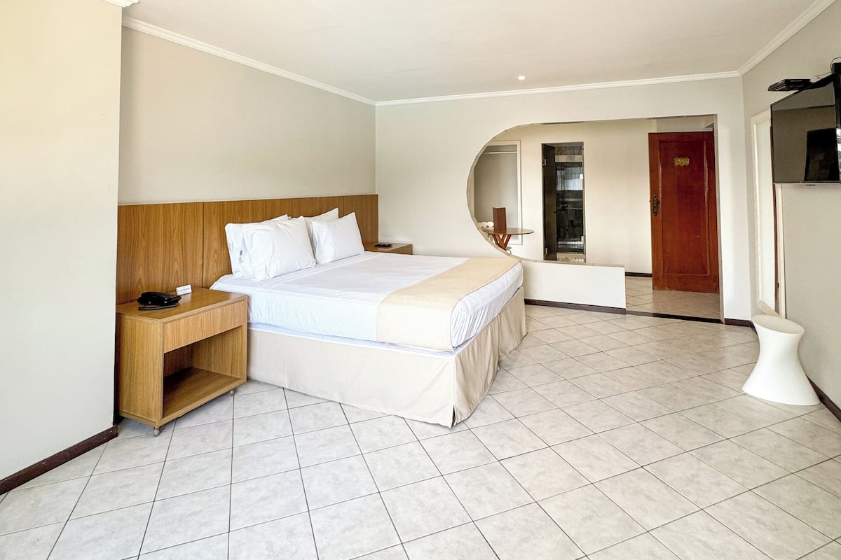 Amplia habitación de hotel con una cama grande, una mesita de noche de madera y un tocador, con un baño visible a través de un arco.