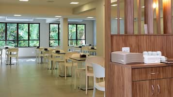 A spacious dining area with multiple tables and chairs, large windows with a view of greenery, and a wooden counter with cups and a tissue box.