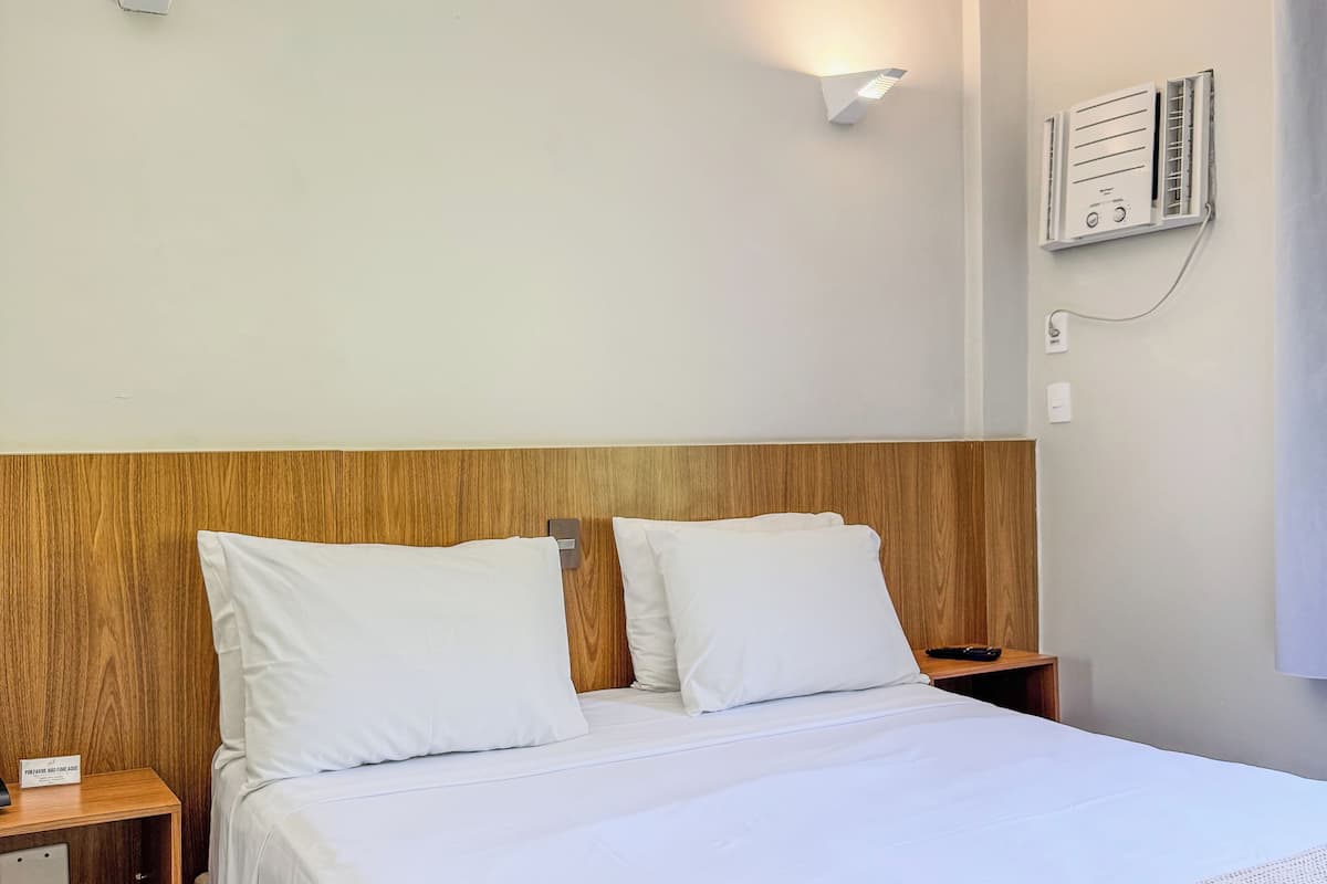 Habitación de hotel con cabecera de madera, ropa de cama blanca, mesita de noche con lámpara y aire acondicionado de pared.